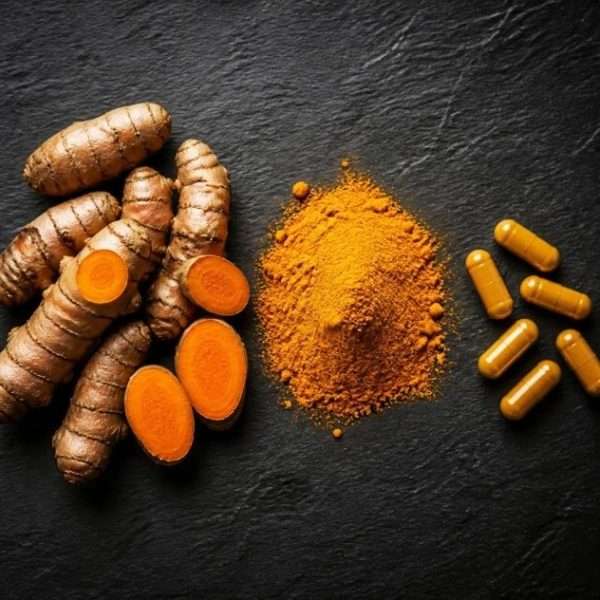 Turmeric roots with sliced pieces, a mound of turmeric powder, and several yellow capsules on a dark surface.